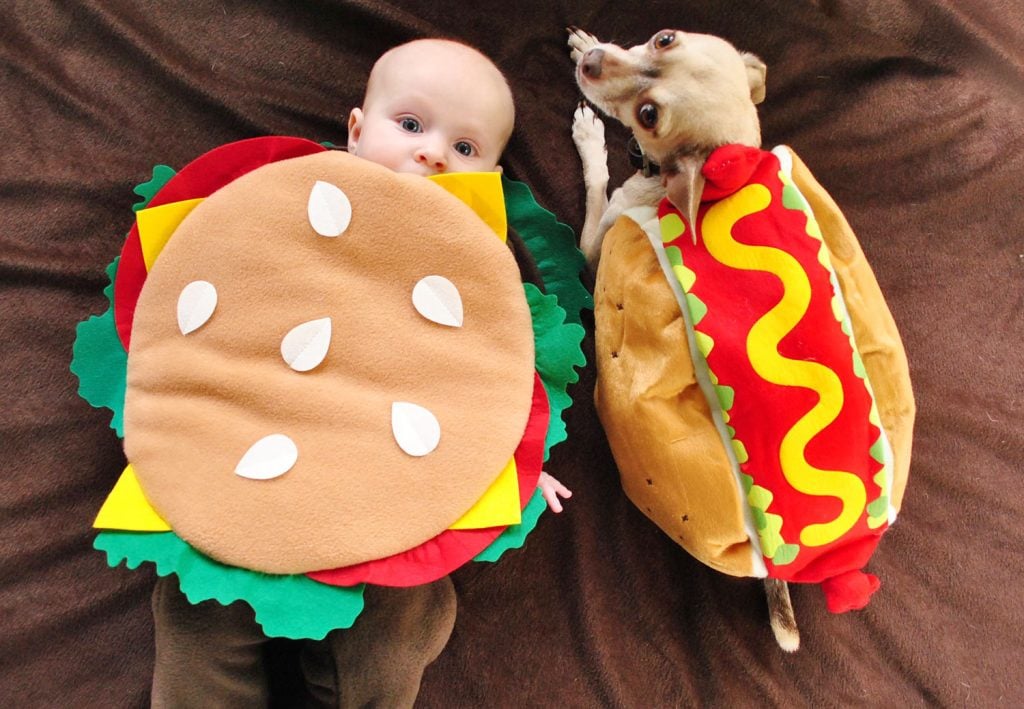 Baby Dressed as Hamburger Next to Dog In Hot Dog Costume